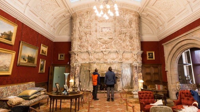 Two visitors are looking up at an enormous, highly decorated marble fireplace in the Drawing Room at Cragside